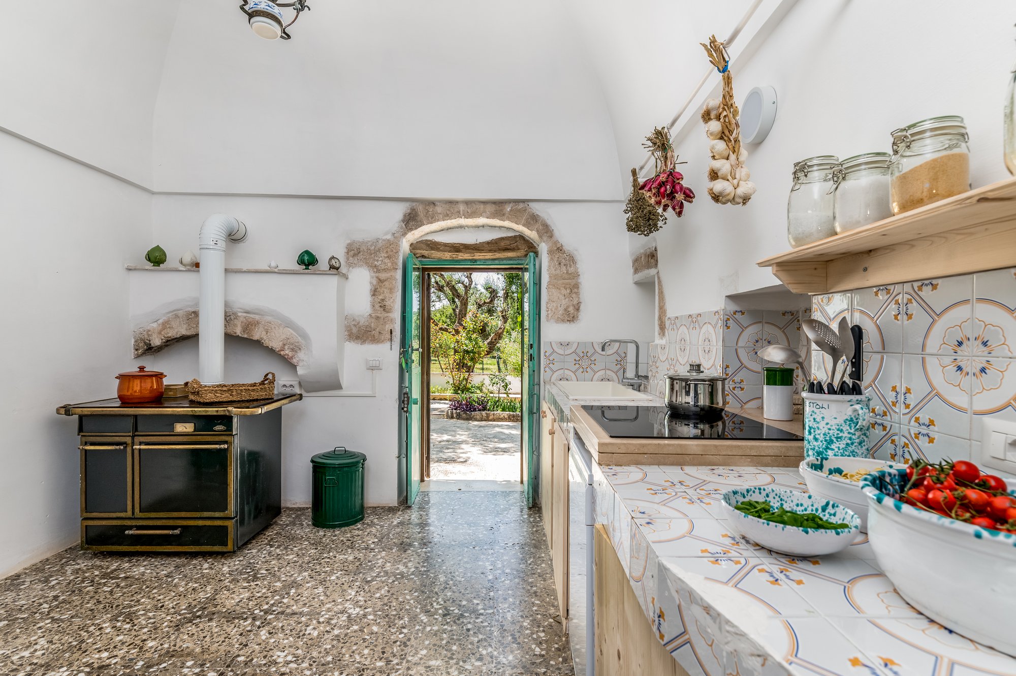 Cucina in pietra con cucina economica, ceramiche pugliesi e porta verde sul giardino della masseria